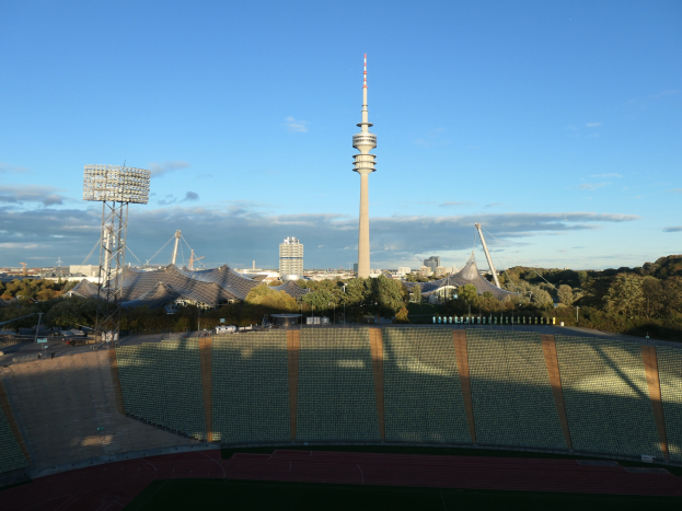 Olympiastadion in Berlin, Deutschland, mit dem Fernsehturm im Hintergrund, umgeben von Bäumen, Gebäuden und beleuchteten Bereichen unter einem bewölkten Himmel.