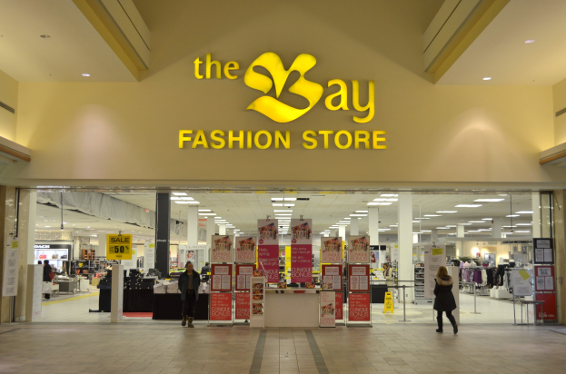 Außenansicht eines Bay Fashion Stores in einem Einkaufszentrum mit Passanten, Wandtextanzeigen, Säulen, Deckenleuchten und Hintergrunddekoration.