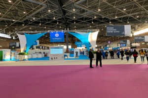 Ein gesch├Ąftiges Messestand auf der Pariser Internationalen Handelsmesse 2018, mit Menschen, die herumlaufen, T├╝ten tragen und Gegenst├Ąnde auf Tischen betrachten, unter h├Ąngenden Bannern und Deckenleuchten.