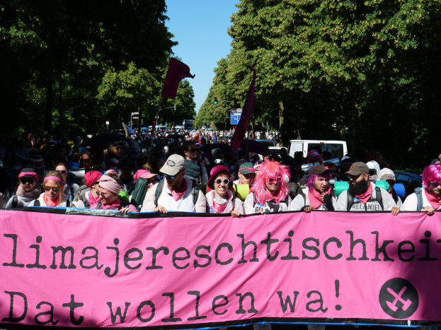 Eine Gruppe von Menschen marschiert die Straße entlang und hölt eine rosa Banner mit der Aufschrift 'Frauenmarsch in Berlin', wobei einige Mönche Kappen und Göggel tragen und andere Fahnen tragen, vor dem Hintergrund von Bäumen, Fahrzeugen und Himmel.