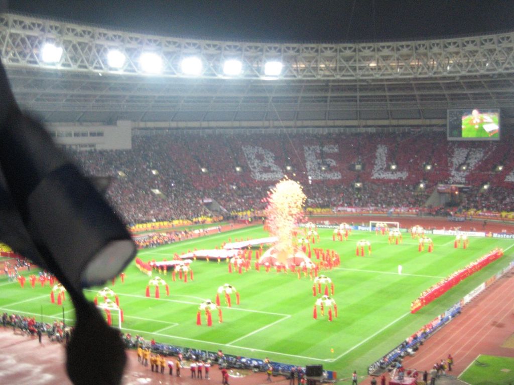 Ein Stadion mit Flutlicht, Eisengrills, Zuschauern, stehenden Individuen, Werbetafeln, Anzeigetafeln, Sicherheitskameras, Flaggen und Dekorationen.