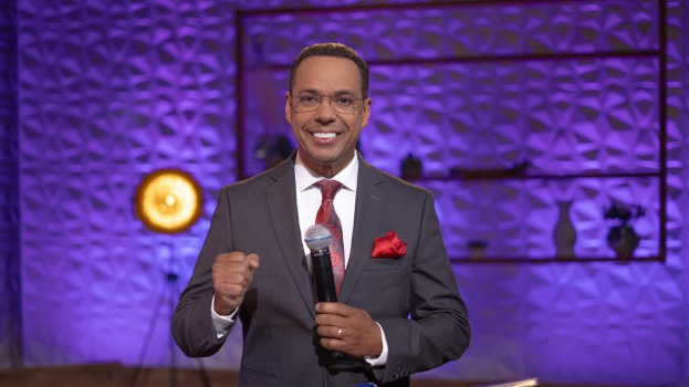 Stephen Colbert, Moderator von 'The Daily Show with Trevor Noah', steht auf einer Bühne und hält ein Mikrofon in der Hand, trägt einen Anzug, Krawatte und Brille, hinter ihm ist ein Scheinwerfer und ein Ständer zu sehen.