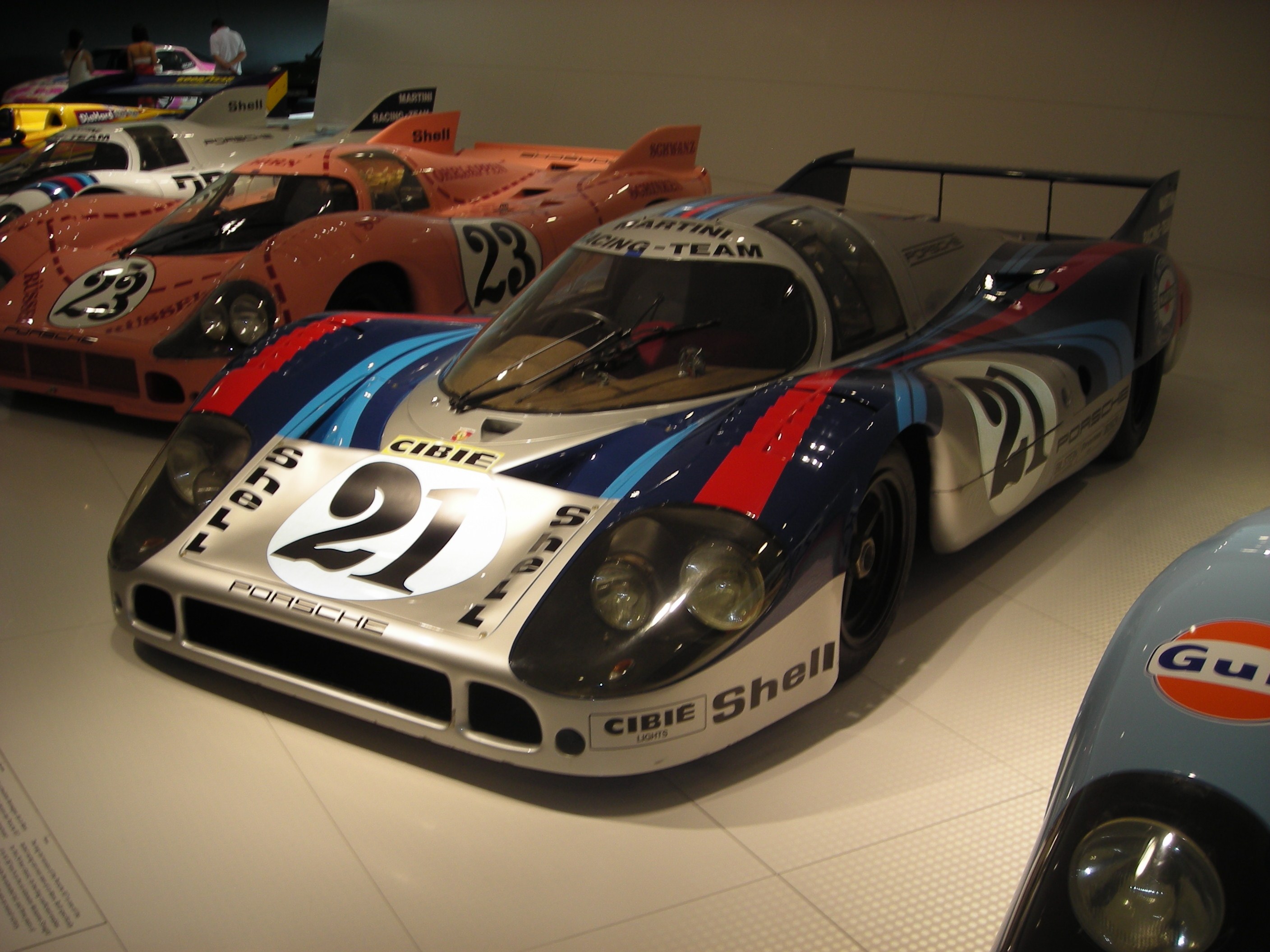 Ein Porsche 917K Le Mans Rennauto, das in einem Museum ausgestellt ist, umgeben von anderen Sportwagen auf dem Boden, mit Menschen in der Nähe und informativem Text auf der linken Seite.