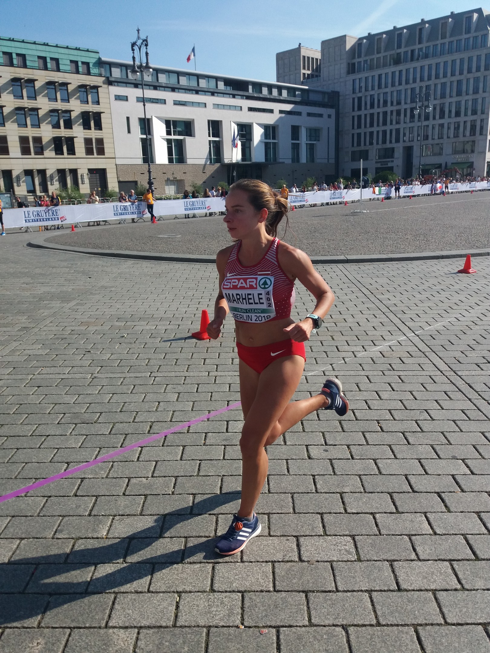 Eine Frau in Sportkleidung läuft bei einem Marathon an Verkehrskegeln, einer Schleife, Zuschauern, einem Banner, Straßenlaternen mit Fahnen, Gebäuden und einem bewölkten Himmel vorbei.