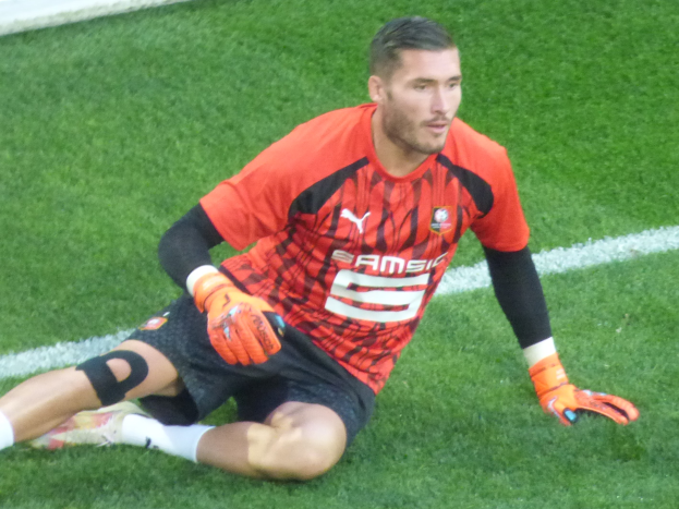 Ein Fußballspieler in rotem T-Shirt, schwarzen Shorts, weißen Socken und orangefarbenen Baseball-Handschuhen sitzt auf dem Boden mit den Händen auf den Knien.