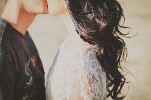 Ein Paar in Hochzeitskleidung umarmt und küsst sich leidenschaftlich bei Sonnenuntergang, wobei die Frau ein weißes Kleid trägt und der Mann ein schwarzes T-Shirt, während im Hintergrund warmes orangefarbenes Licht leuchtet.