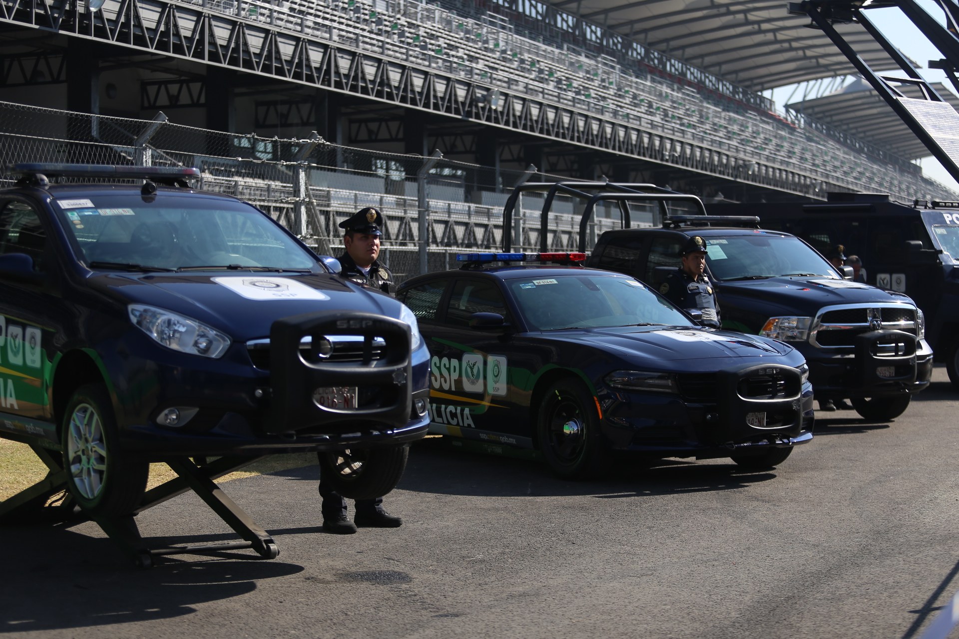 Polizeiautos vor einer Rennstrecke geparkt, uniformierte Personen auf der Straße stehend, Stadion und Maschendrahtzaun im Hintergrund.