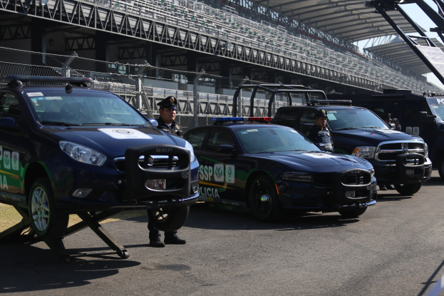 Polizeiautos vor einer Rennstrecke geparkt, uniformierte Personen auf der Straße stehend, Stadion und Maschendrahtzaun im Hintergrund.