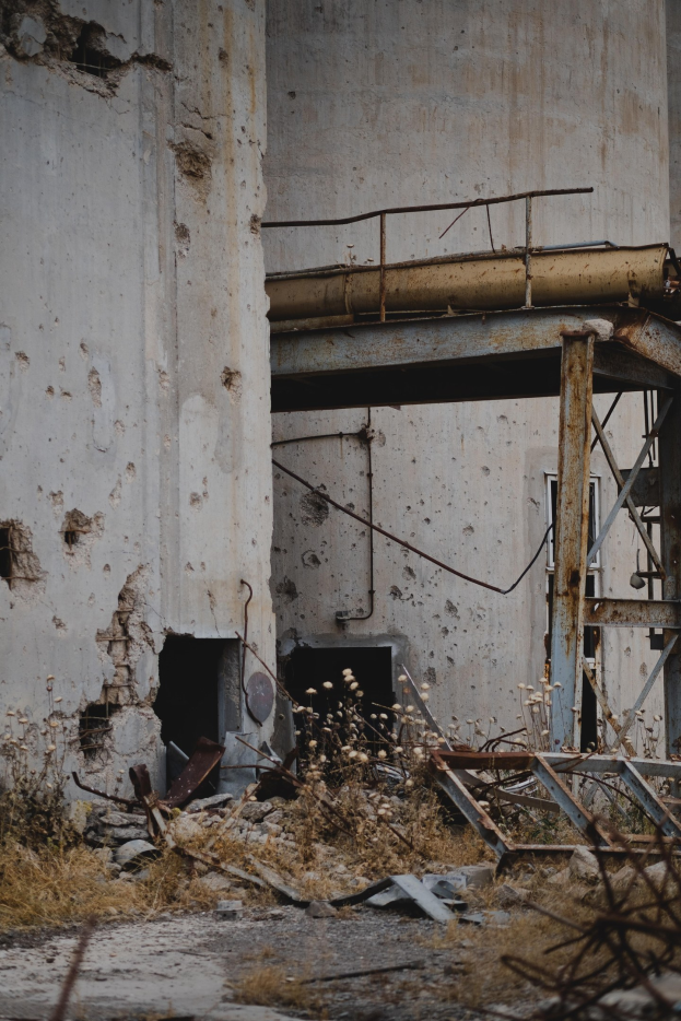 Altes Gebäude mit einem zentralen Metallgerüst, umgeben von Pflanzen, Eisenstangen und Schutt, mit bröckelnden Wänden und zerbrochenen Fenstern.