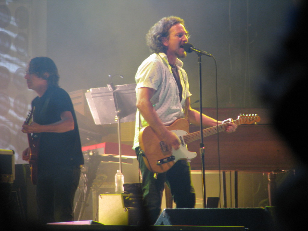 Ein Mann in einem weißen Hemd spielt Gitarre und singt in ein Mikrofon auf der Bühne, neben ihm spielt ein anderer Mann in einem schwarzen Hemd Gitarre; ein Piano, Kabel, Lautsprecher und Lichter sind im Hintergrund zu sehen.