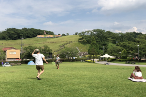Menschen spielen Badminton in einem Park mit Zelten, Straßenmöbeln und Gebäuden im Hintergrund.