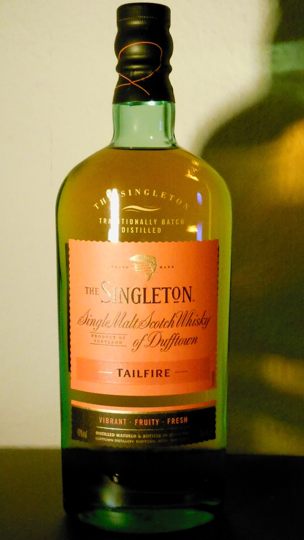 Eine tiefe bernsteinfarbene Flasche Singleton of Dufftown Tailfire Single Malt Scotch Whisky auf einem Tisch, mit einem schwarzen Etikett mit fetter Schrift und einem Wappen mit einem Löwen und einem Schloss.