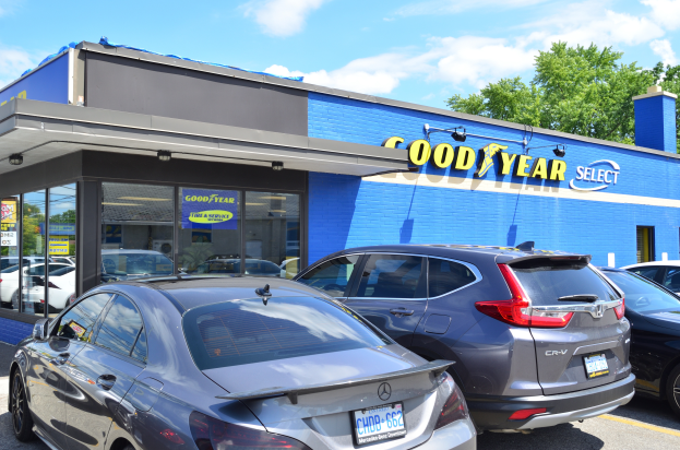 Ein Goodyear Select Autohaus mit mehreren geparkten Autos davor, das einen Text auf dem Gebäude und einen Hintergrund mit Bäumen unter einem bewölkten Himmel zeigt.