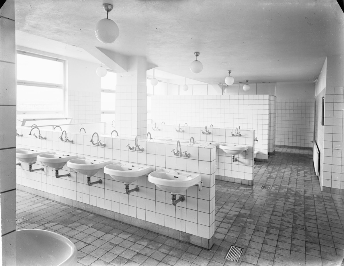 Schwarz-weiß-Foto einer öffentlichen Toilette mit Waschbecken, Hähnen, Wänden, Lampen und Fenstern.