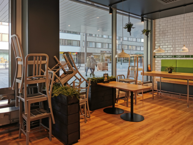 Ein Restaurant in Berlin mit Holztischen und -stühlen, das durch ein großes Fenster zu sehen ist, umgeben von Pflanzen und Beleuchtung, mit Passanten und Gebäuden draußen.