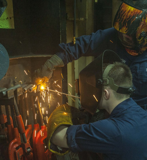 Zwei Schweißer in Schutzausrüstung arbeiten an einem Metallstück in einer Fabrik, einer hält einen Schweißbrenner, mit Werkzeugen und Equipment im Hintergrund.