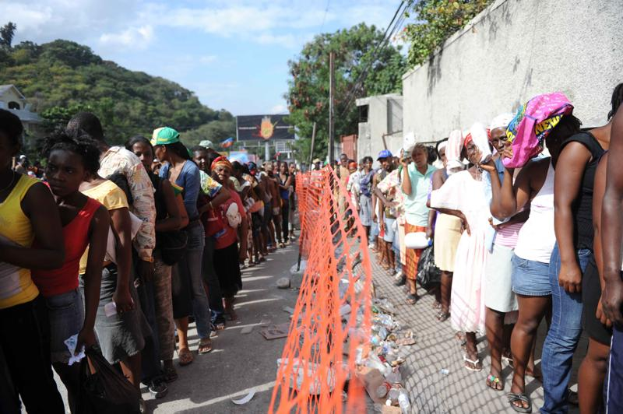 Eine lange Schlange von Menschen steht vor einem Zaun, mit einer Wand auf der rechten Seite, die auf die Stimmabgabe an einer Wahllokal wartet; im Hintergrund gibt es Bäume, Gebäude, Pfosten, Drähte und eine Wand, und der Himmel ist oben sichtbar.
