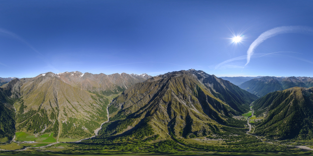 Panoramischer Blick auf ein Gebirgsmassiv von einem Hügel aus, mit grünem Gras, einer gewundenen Straße und einem hellblauen Himmel mit Sonnenlicht.