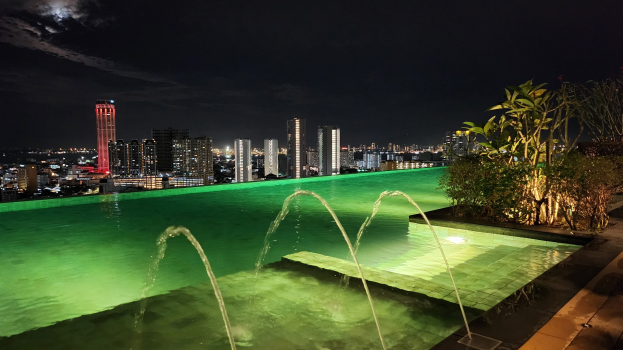 Ein Schwimmbad mit einer zentralen Fontäne, umgeben von Pflanzen auf der rechten Seite und Gebäuden im Hintergrund, beleuchtet von Stadtlichtern in der Nacht.