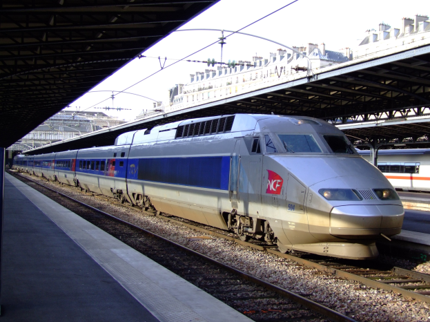 Schnellzug fährt auf Schienen neben einem Bahnsteig mit Gebäuden und einem klaren blauen Himmel im Hintergrund.