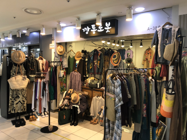 Kleidungsgeschäft mit Kleiderstangen, Hüten, Taschen und Schuhen, beleuchtet von Deckenlampen und mit Texttafeln.