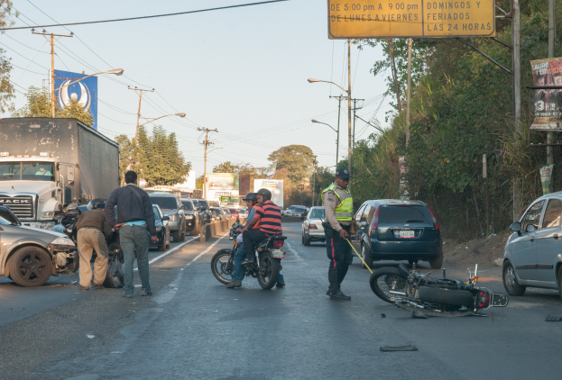 Gruppe von Menschen, die um ein verunglücktes Motorrad auf der Seite einer Straße stehen, mit Fahrzeugen, Bäumen, Masten und Lichtern im Hintergrund.