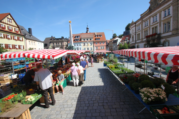 Ein belebter Markt im historischen Stadtkern von Heidelberg, Deutschland, mit Menschen, die spazieren gehen, auf Bänken sitzen und in der Nähe von Zelten, Tischen mit Körben voller Gemüse und Gebäuden mit Fenstern, Bäumen und einem klaren blauen Himmel im Hintergrund.