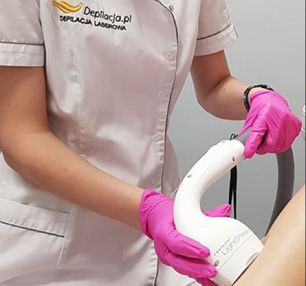 Eine Frau in einem weißen Hemd und pinken Handschuhen verwendet eine weiße Laser-Haarentfernungsmaschine auf ihrer Haut, während sie an einer Wand steht.