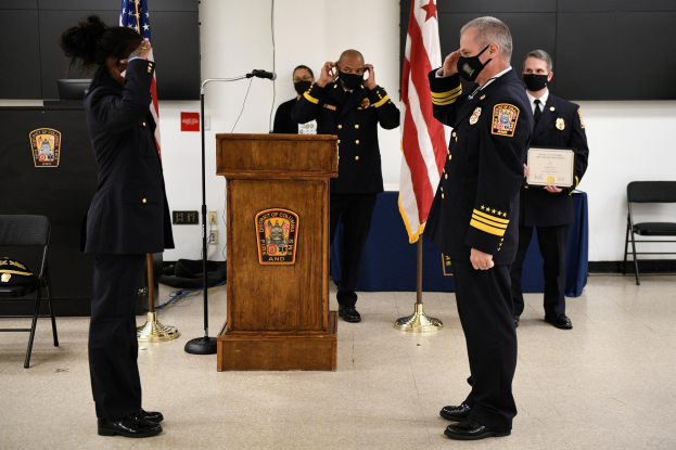 Gruppe uniformierter Personen in Masken vor einem Podium mit Mikrofon, umgeben von Fahnen und Möbeln, bei einer Vereidigung eines neuen Feuerwehrchefs.