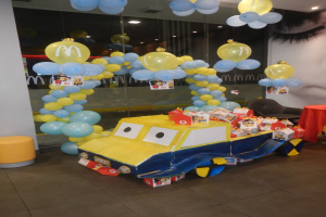 Eine McDonald's-Geburtstagsfeier mit einem gelben und blauen Lastwagen, der mit Ballons und anderen Dekorationen geschmückt ist, einem Tisch mit roter Decke und verschiedenen Gegenständen darauf, Hockern, einer Glaswand und einer mit Lichtern geschmückten Decke, die eine festliche Atmosphäre schaffen.