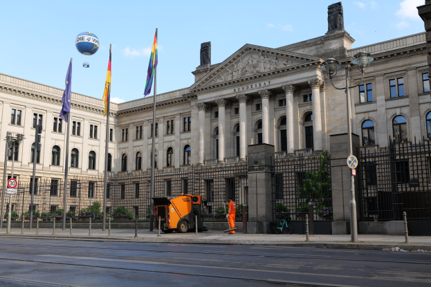Großes modernes Regierungsgebäude (Bundestag, Berlin) mit vielen Fenstern, umgeben von Straßeninfrastruktur, Fußgängern, Flaggen und Bäumen unter einem bewölkten Himmel.