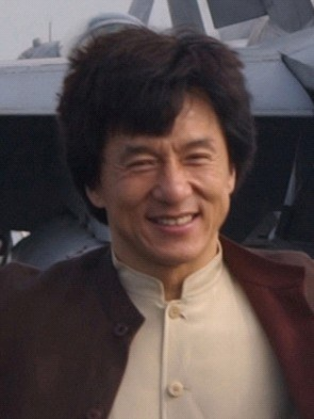 Jackie Chan steht lachend vor einem Flugzeug gegen einen klaren blauen Himmel, trägt ein weißes Hemd, eine schwarze Jacke und hat ordentlich frisiertes Haar.