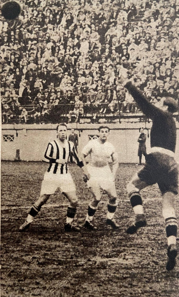 Schwarz-weißes Foto von Männern, die Füßball spielen, mit Zuschauern auf der Tribüne und einer Wand im Hintergrund, beschriftet mit "Newcastle United v West Bromwich Albion".