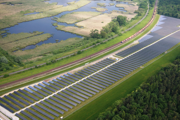Luftaufnahme einer Solarpark mit Panelen, umgeben von Bäumen, Gras, Wasser und einer Bahn auf einem Bahngleis.