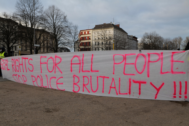 Eine Gruppe von Menschen, die auf dem Boden stehen und ein Banner halten, auf dem "Rechte für alle Menschen Stoppt Polizeigewalt" steht, mit einem Straßenschild, einem Schild, Bäumen, Gebäuden mit Fenstern und einem bewölkten Himmel im Hintergrund.