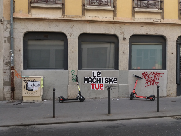Zwei Roller auf einer Straße neben einem Gebäude mit Fenstern, Geländern und einem Rohr geparkt, mit Masten und einer Texttafel in der Nähe.