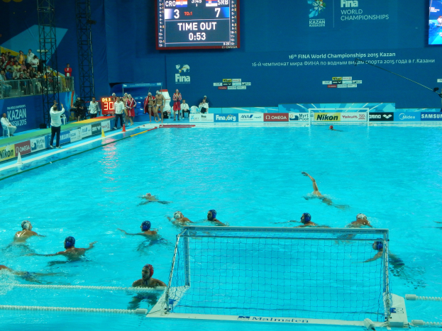 Eine Gruppe von Menschen spielt Wasserball in einem Pool mit einem Netz, umgeben von Zuschauern, Tafeln mit Text und Logos, Trägern und einem Bildschirm mit Zahlen und Text bei den 2015 FINA World Championships in Kazan.
