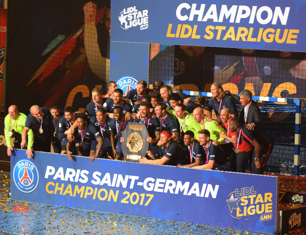 Spieler von Paris Saint-Germain feiern mit der Ligue-1-Trophäe, tragen Medaillen, vor einer Tafel mit Text und Logos, mit einem Netz auf der rechten Seite und einem Bildschirm im Hintergrund.