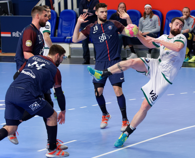 Eine Gruppe von Männern spielt Handball auf einem Platz, wobei einer den Ball hält, während Zuschauer von Stühlen, Geländern und einer Wand im Hintergrund zusehen.