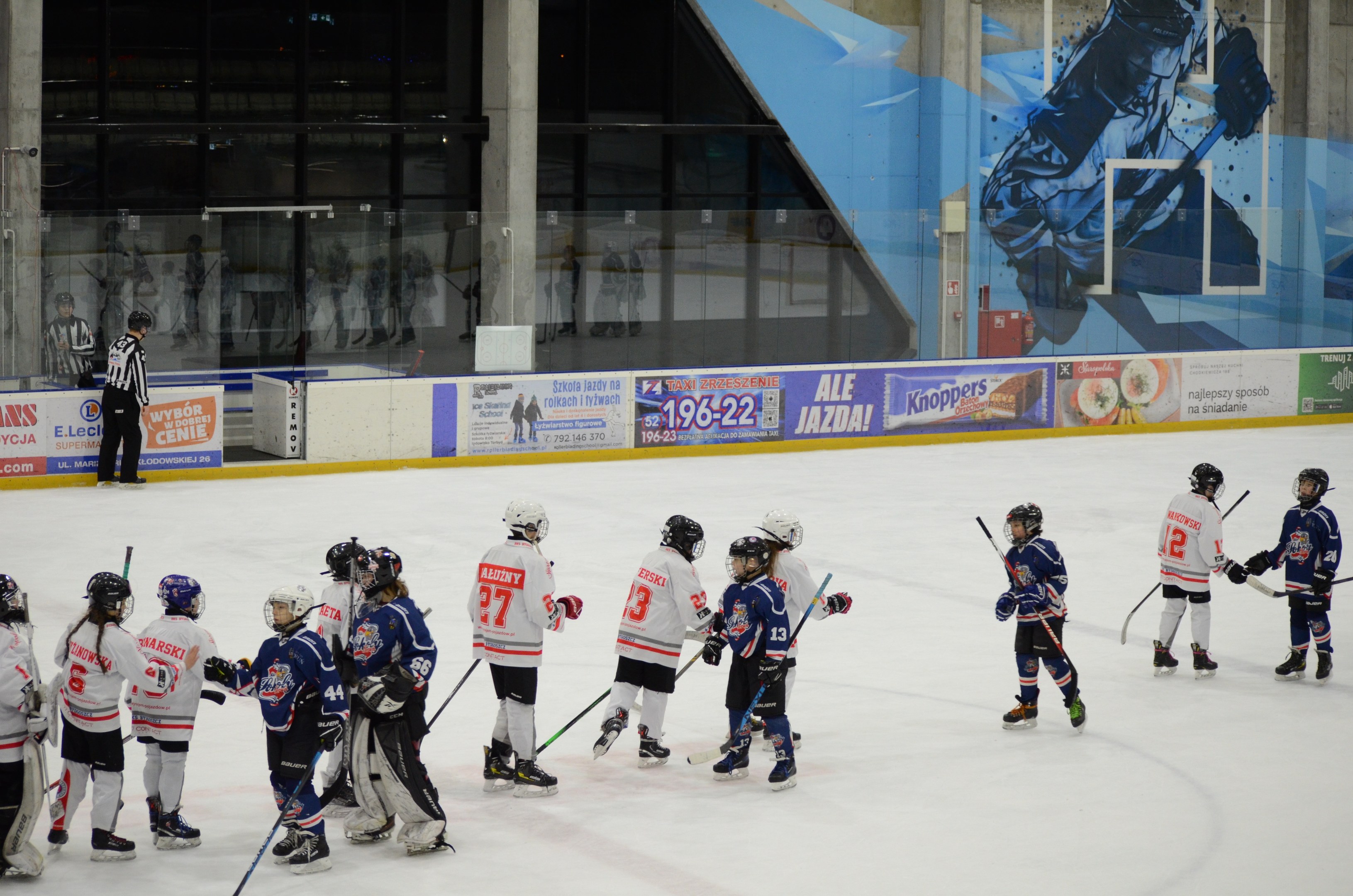 Eine Gruppe junger Menschen, die Eishockey auf einer Indoor-Eisfläche spielen, tragen Helme und halten Stöcke, mit einer Person links daneben und Bannern, Säulen und Glaswänden im Hintergrund.