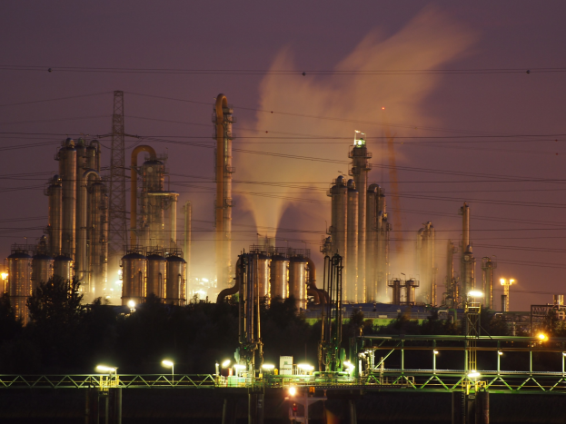 Ölraffinerie bei Nacht mit Rauch aus Schloten, beleuchtet von nahen Lichtern, vor einem sichtbaren Himmel, mit einem Wasserzeichen.