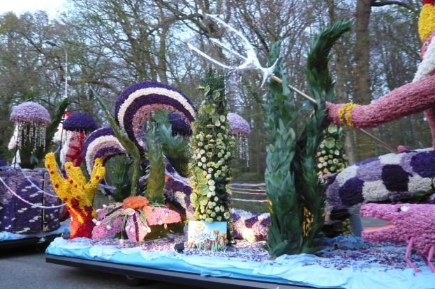 Ein Paradewagen, der mit einer Vielzahl farbenfroher Blumen wie Rosen, Gänseblümchen und Lilien geschmückt ist, umgeben von Bäumen unter einem klaren blauen Himmel.