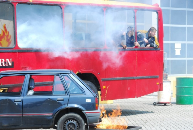 Ein roter Doppeldeckerbus mit Rauch, der aus ihm austritt, mit drei sichtbaren Passagieren, neben einem Auto geparkt, vor einem Gebäude mit Glasfenstern und einem Fass auf der rechten Seite.