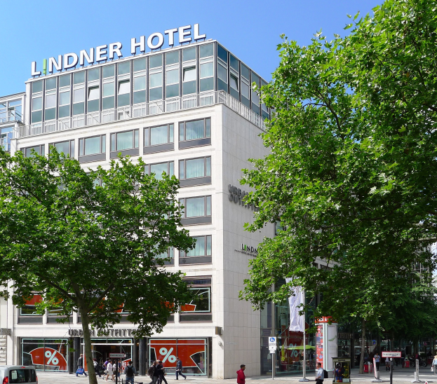 Großes modernes Hotelgebäude mit Glasfenstern, der Name "Lindner" oben drauf, umgeben von Bäumen und einer belebten Straße mit Fußgängern und Fahrzeugen.