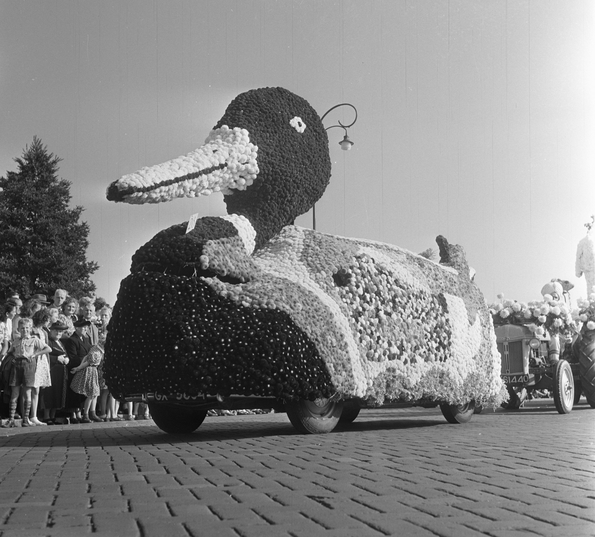 Schwarzes und weißes Foto eines Entenparade-Wagen, der mit Blumen geschmückt ist, mit Menschen zu seiner Linken und Bäumen im Hintergrund unter einem klaren Himmel.