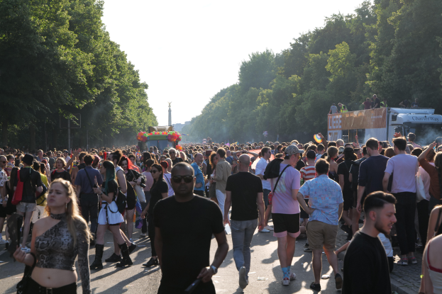 Eine große Menschenmenge, die eine von Bäumen gesäumte Straße mit einem Turm im Hintergrund und Fahrzeugen mit Teilnehmern auf der rechten Seite entlanggeht, wahrscheinlich während des Christopher Street Day in Berlin.