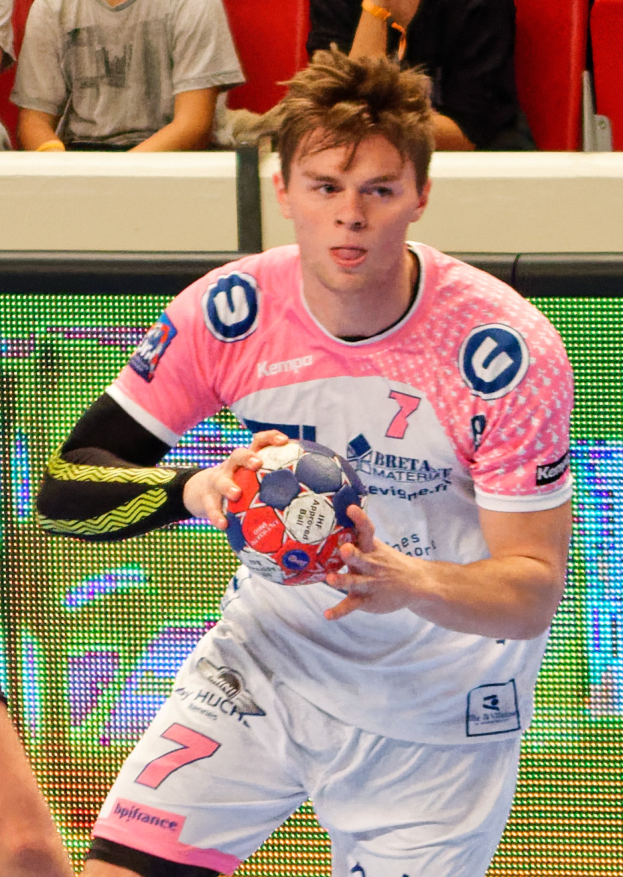 Ein Mann in einem pinken Hemd hält einen Fußball, daneben ist die Hand einer anderen Person zu sehen, im Hintergrund sitzen Menschen auf Stühlen.