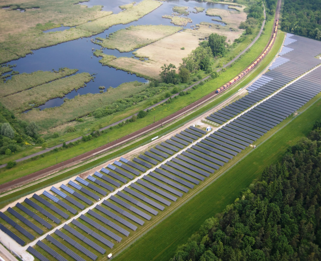 Luftaufnahme eines Solarparks mit Solarpanelen in einem Feld, umgeben von Bäumen, Gras, Pflanzen und Wasser, mit einem Zug auf einer nahen Eisenbahnschiene.