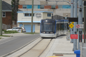 Eine Straßenbahn fährt eine Straße mit hohen Gebäuden entlang, mit Masten, Schildern, einem Mülleimer, einem Auto und einem Baum.