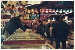 Eine Gruppe von Menschen um einen Faschingsstand mit einer Glasplatte in der Mitte, Spielzeuge hängen an einem Pfahl links und Lichter und andere Gegenstände im Hintergrund.